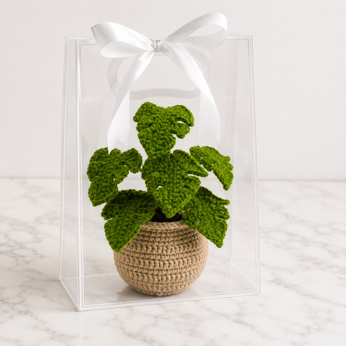 Crochet monstera plant in tan pot inside clear acrylic gift box with white bow