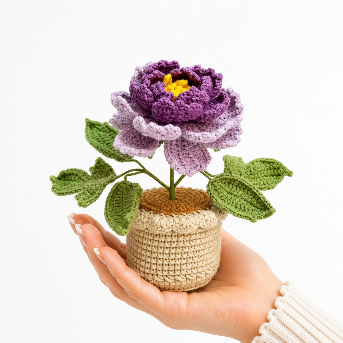 Handmade crochet peony in beige pot held in a hand, purple flower with green leaves