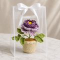 Handmade crochet peony in beige pot with white ribbon bow, purple flower and green leaves