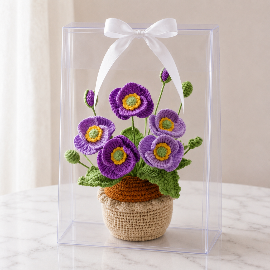 Crocheted purple poppies in beige yarn pot inside clear gift box with white bow