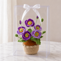 Crocheted purple poppies in beige yarn pot inside clear gift box with white bow