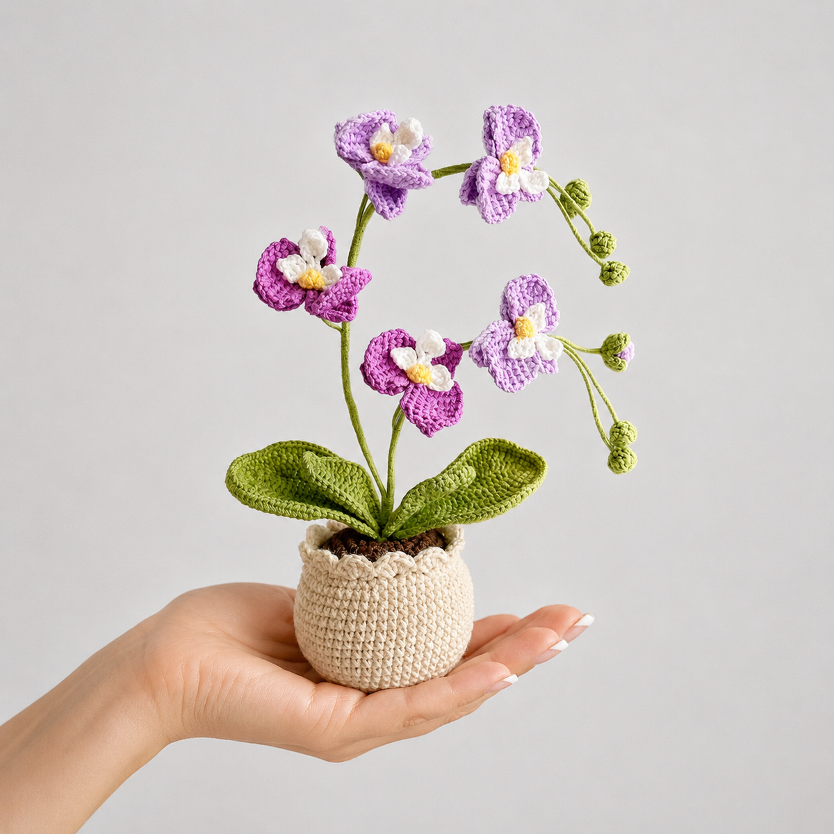 Crocheted purple orchid with green leaves in beige pot, held in a hand