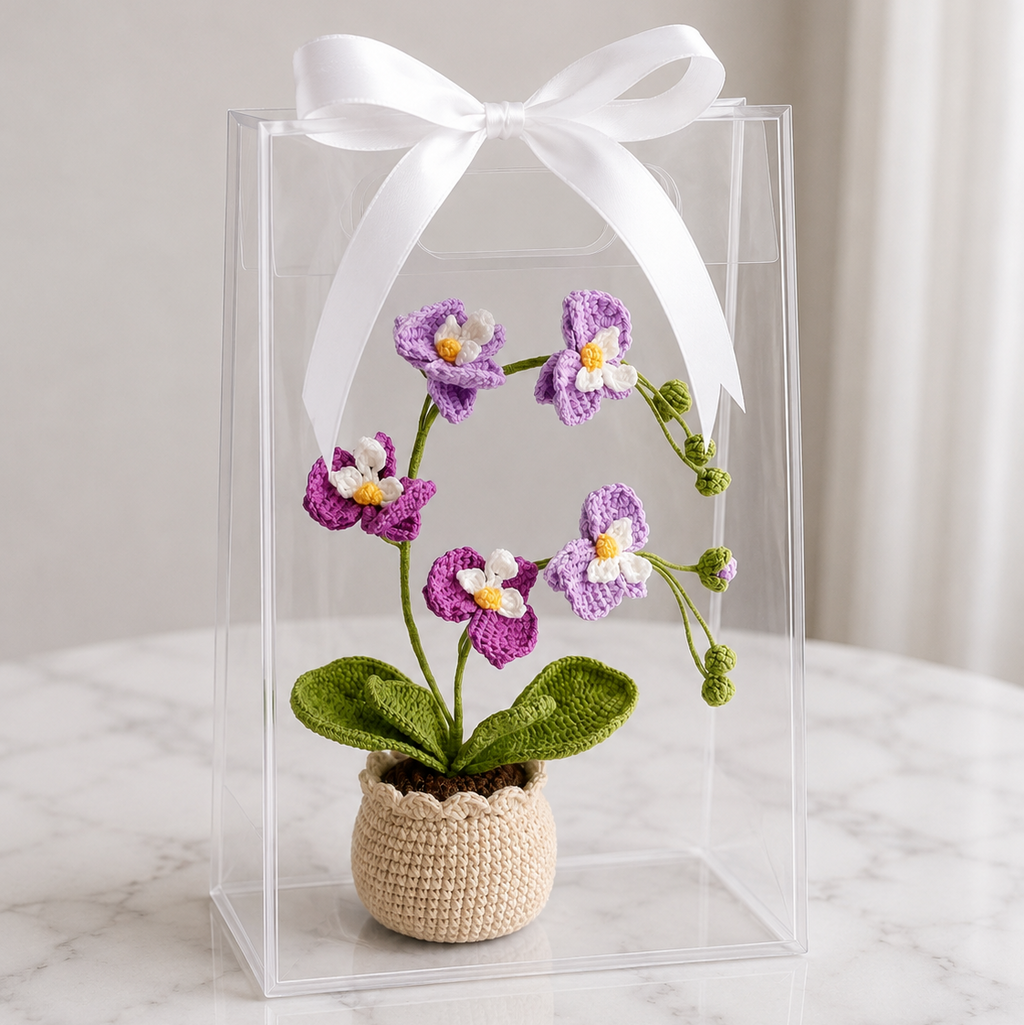 Crocheted purple orchid with green leaves in beige pot, inside clear gift box