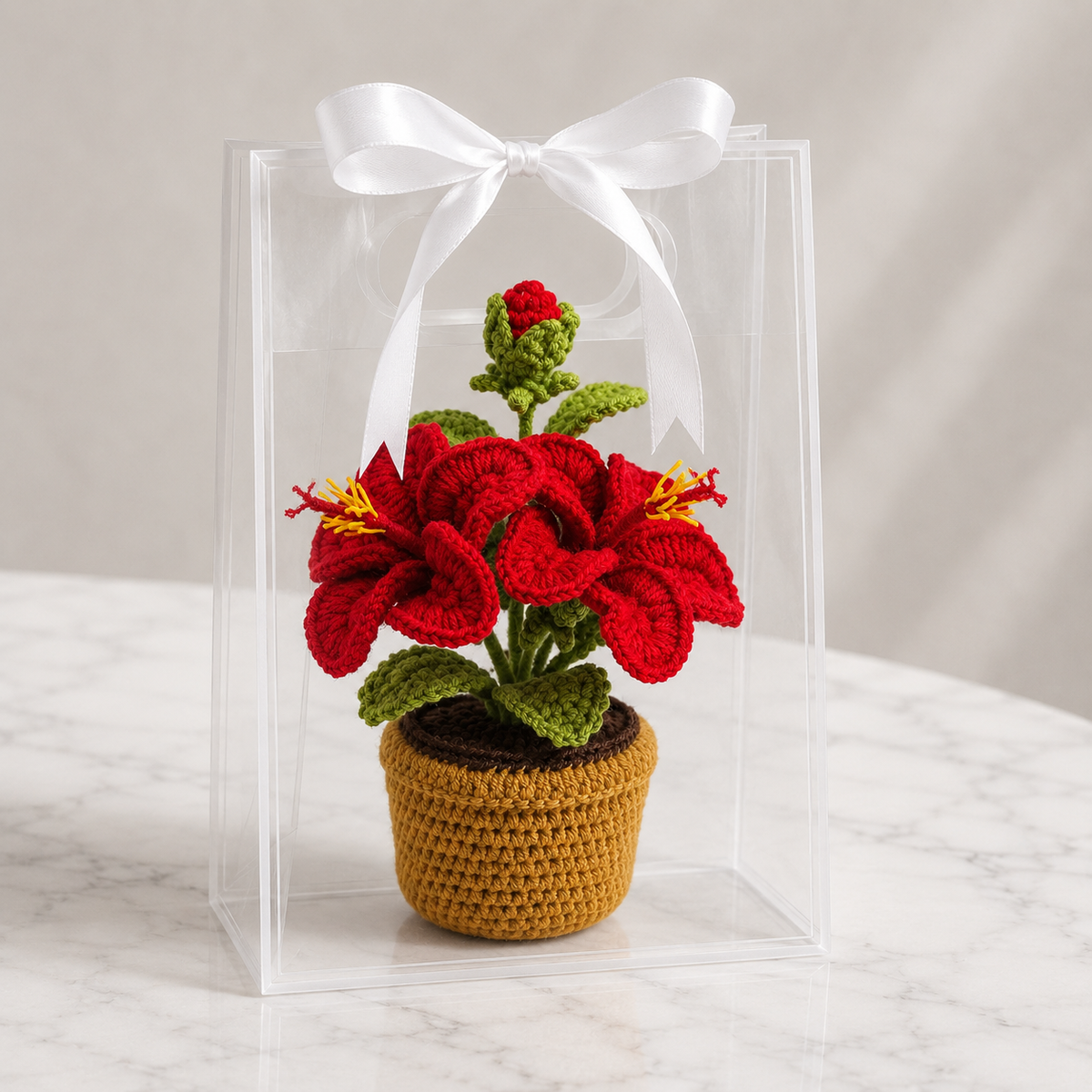 Crocheted red lily amigurumi flower in tan pot, inside clear display box with white ribbon.